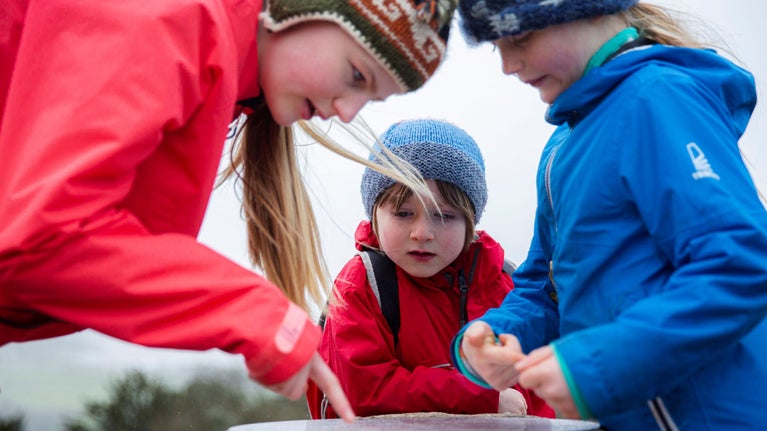 Three children looking at the Pepperpot monument at Arnside and Silverdale. They are all wearing brightly coloured waterproof coats and wooly hats.